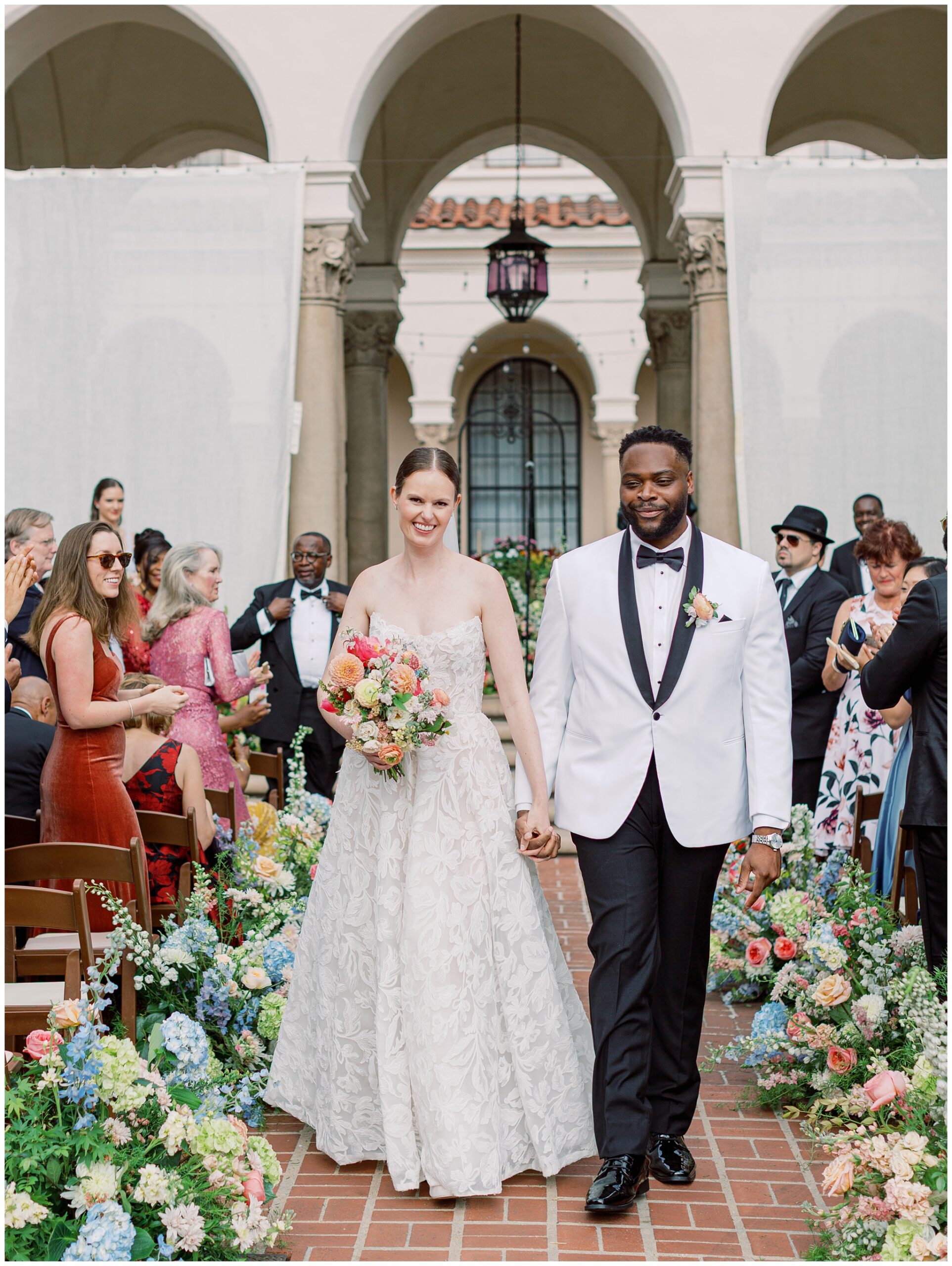 Newlywed couple walking back down the aisle during their Caltech Athenaeum courtyard ceremony as guests smile and colorful floral arrangements surround the space