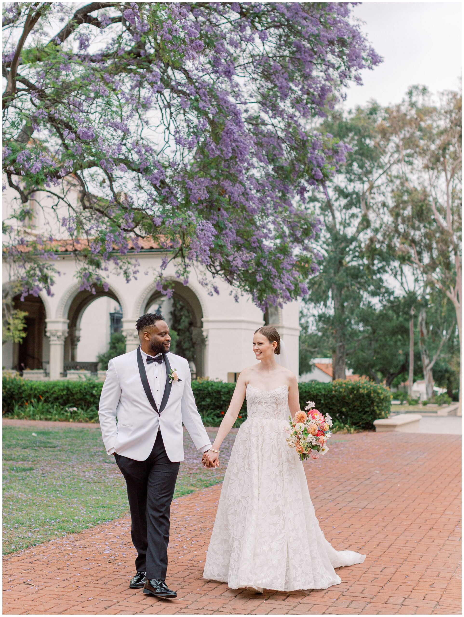 Bride and groom walking beneath the jacaranda tree at the Caltech Athenaeum during their wedding portraits