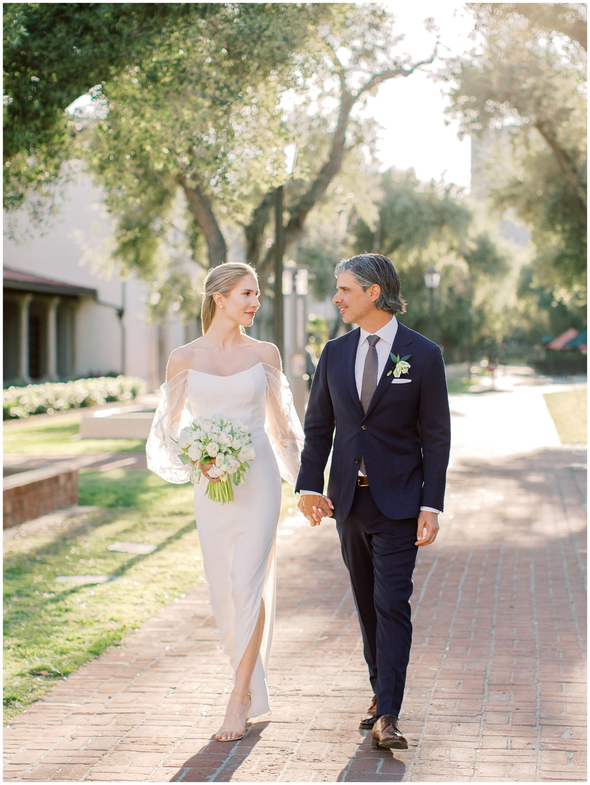 Bride and groom walking hand in hand through the olive tree-lined walkway during their wedding portraits