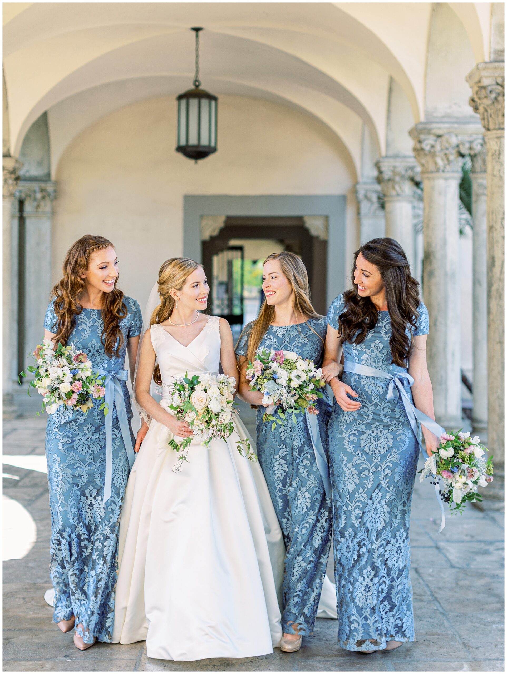 Bride and bridesmaids in blue dresses walking beneath the columns during their wedding portraits