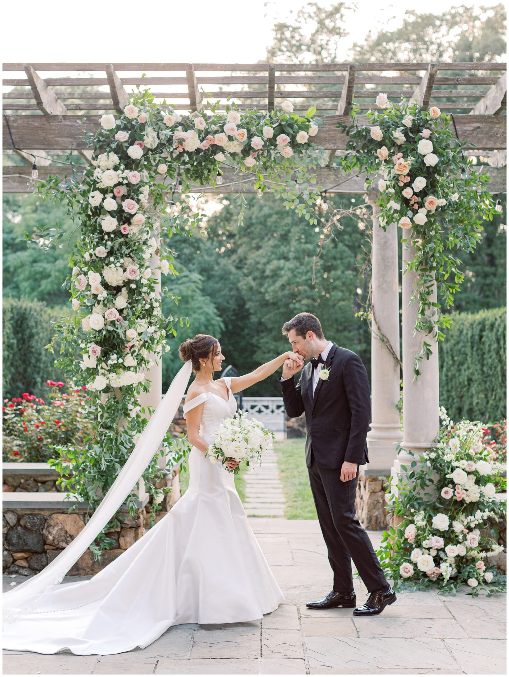 Groom kissing the bride's hand beneath a lush floral ceremony arch on the Bluestone Terrace at Dover Hall