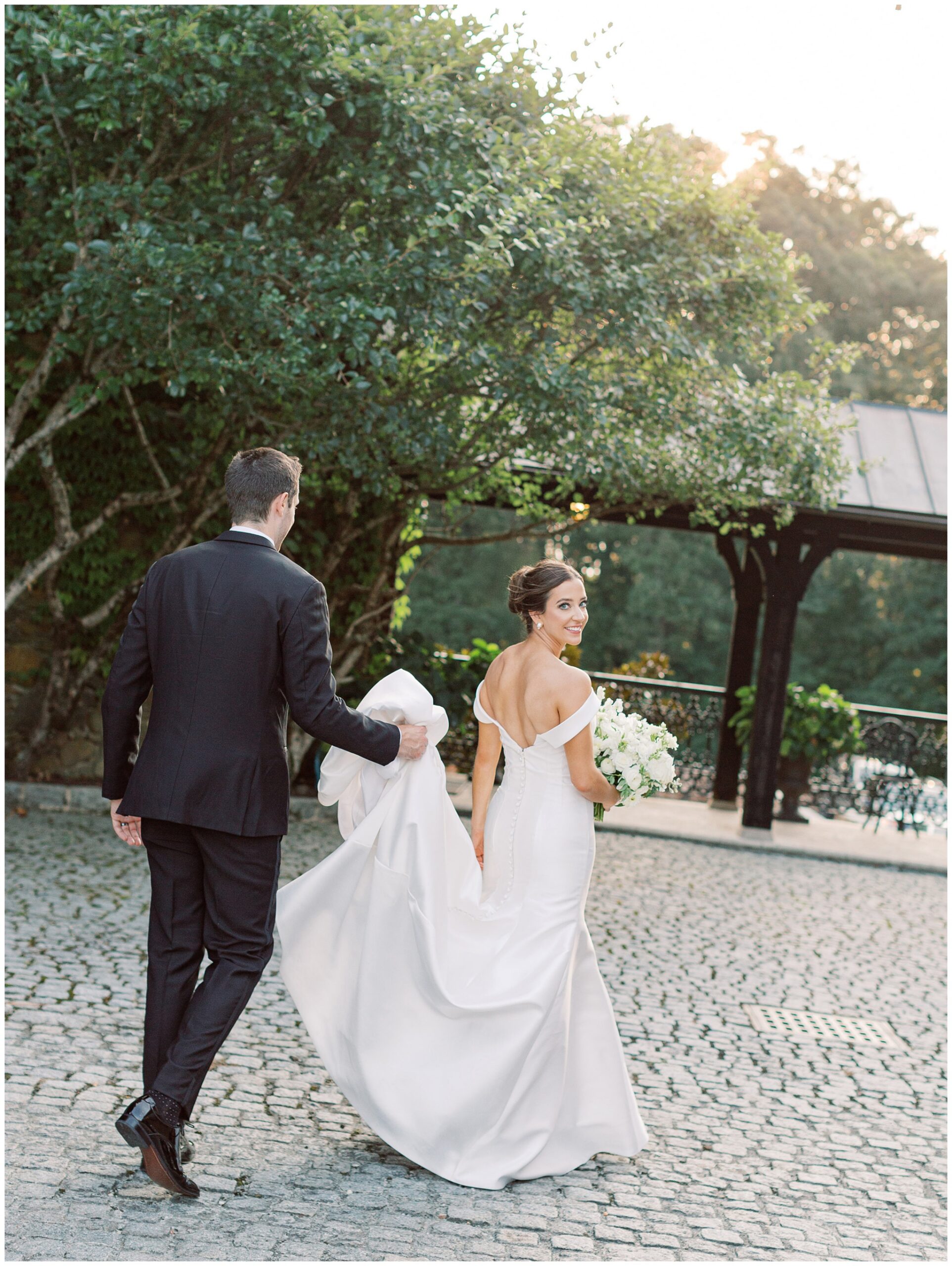 Bride and groom walking through the cobblestone court with the bride looking over her shoulder and groom carrying her wedding dress