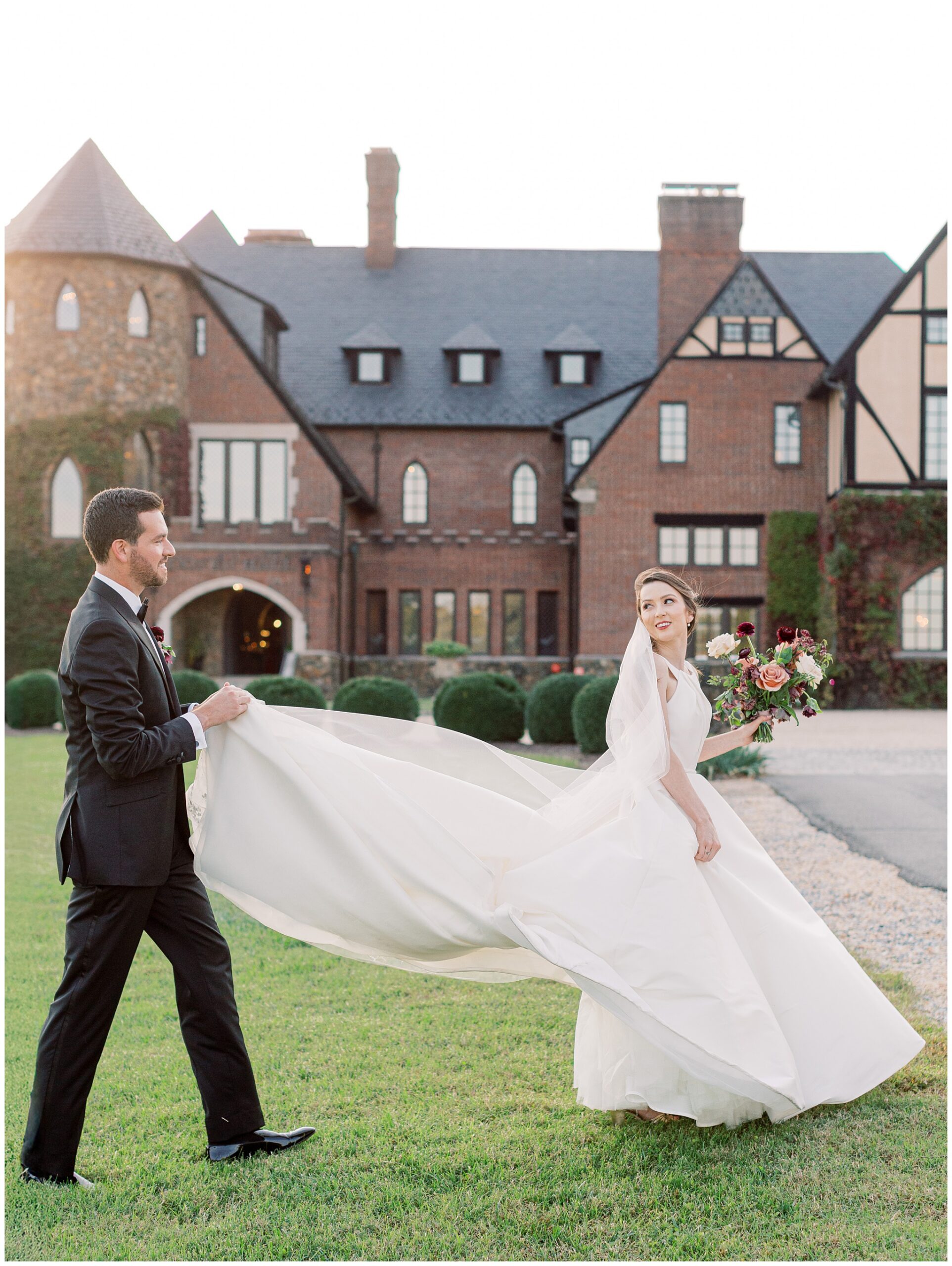 Newlyweds walking across the lawn with the groom carrying the bride’s wedding dress