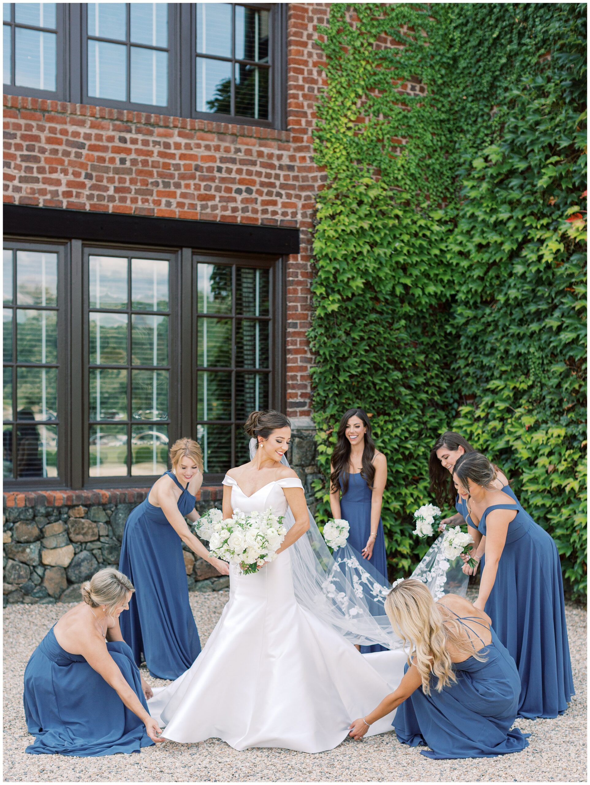 Bride with her bridesmaids adjusting her wedding dress in front of Dover Hall’s ivy-covered exterior