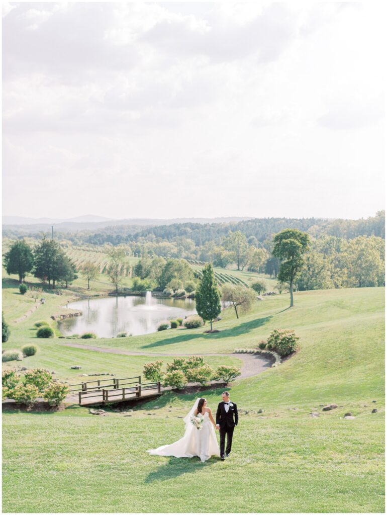 Bride and groom walking near the Stone Circle at Stone Tower Winery with the pond, hydrangeas, and Blue Ridge Mountains in the background